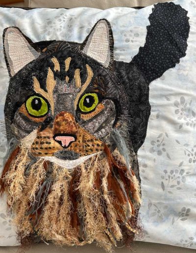 Photo is of fabric cat pet portrait pillow. Cat has dark grey ginger face with yarn to represent its fluffy chest fur.