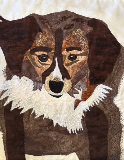 Fabric wallhanging is of a brown dog with dark brown ears and white fluffy fur around the front of its neck. Brown thread is sewn on to indicate the fluffiness of the fur. .